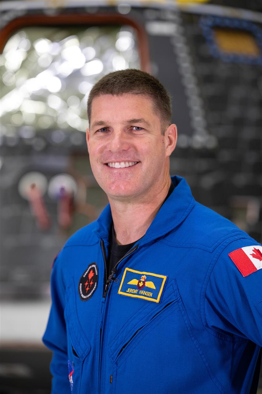 A Canadian astronaut wearing a blue flight suit smiles in front of a tear-shaped capsule.