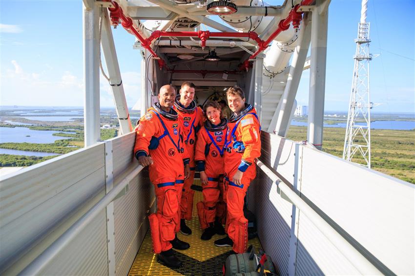 Four astronauts in orange spacesuits pose on a metal walkway high in the air.