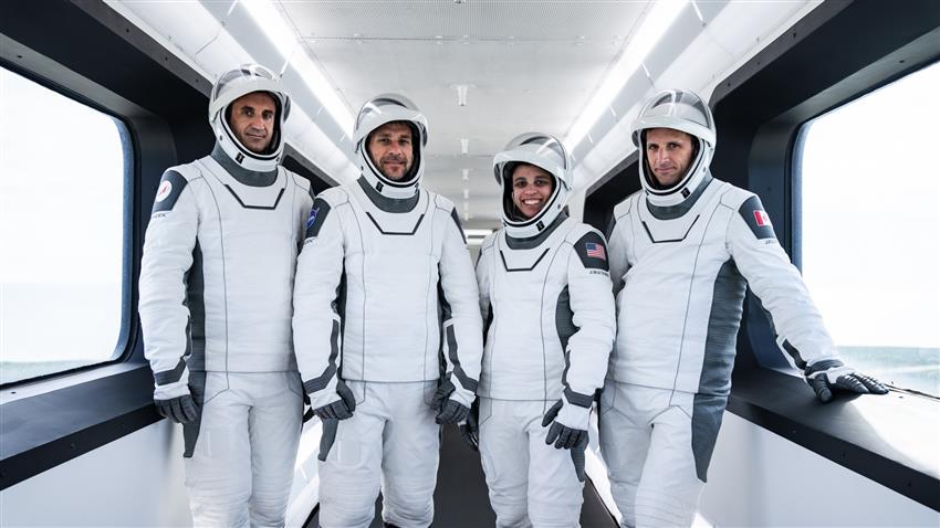 The four astronauts, wearing space suits, stand in a line. 