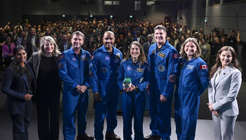 The group poses on stage for the photo. Behind them is a full audience.