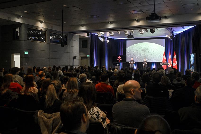 Audience members are seated in a conference room. They are facing a big screen and two people standing on stage.