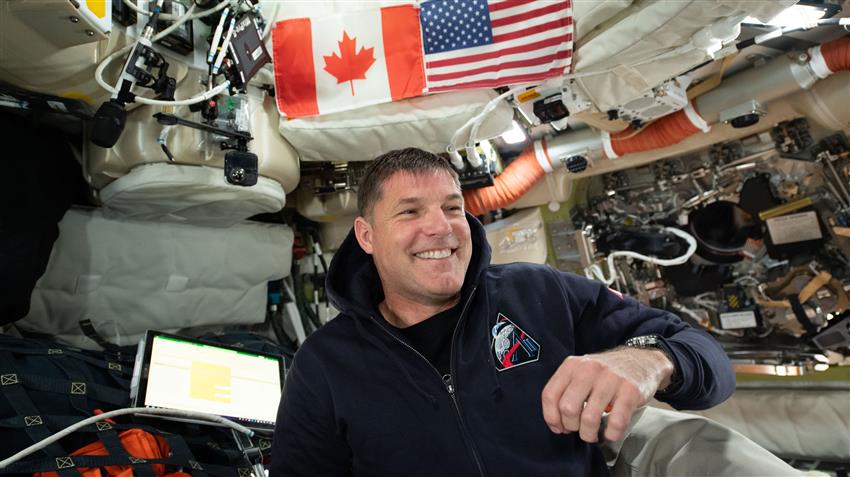 Jeremy sourit en regardant hors caméra. Des drapeaux du Canada et des États-Unis se trouvent derrière lui. 