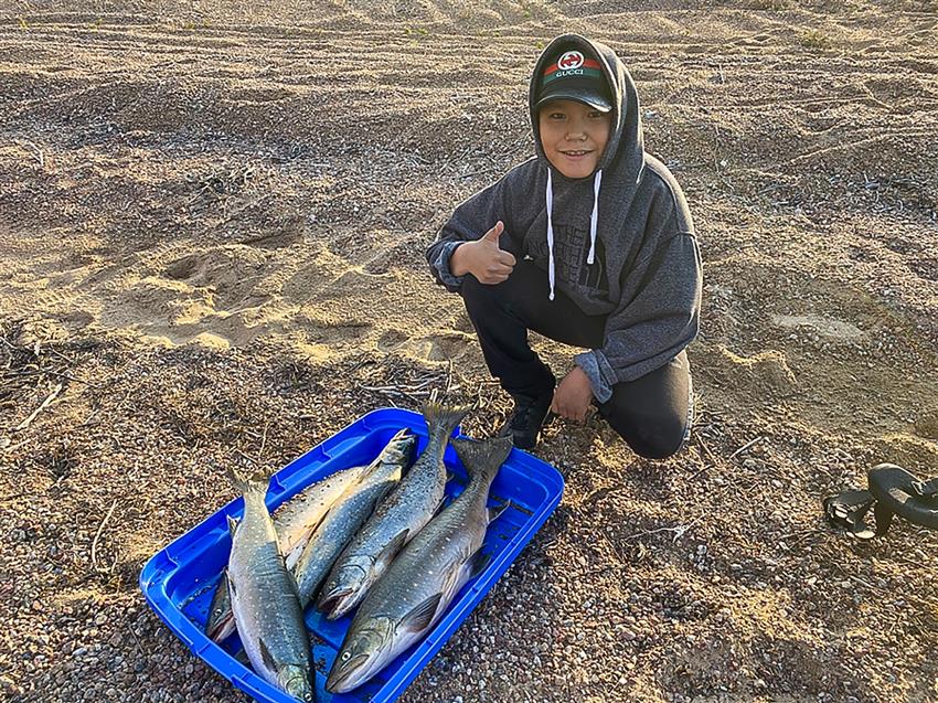 An Indigenous child kneeling in front of a container with five big fish gives a thumbs-up.