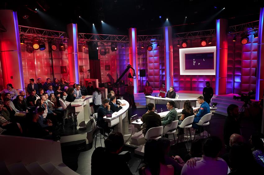 Ten guests and a host of a televised talk show sitting at different desks. They are surrounded by an audience.