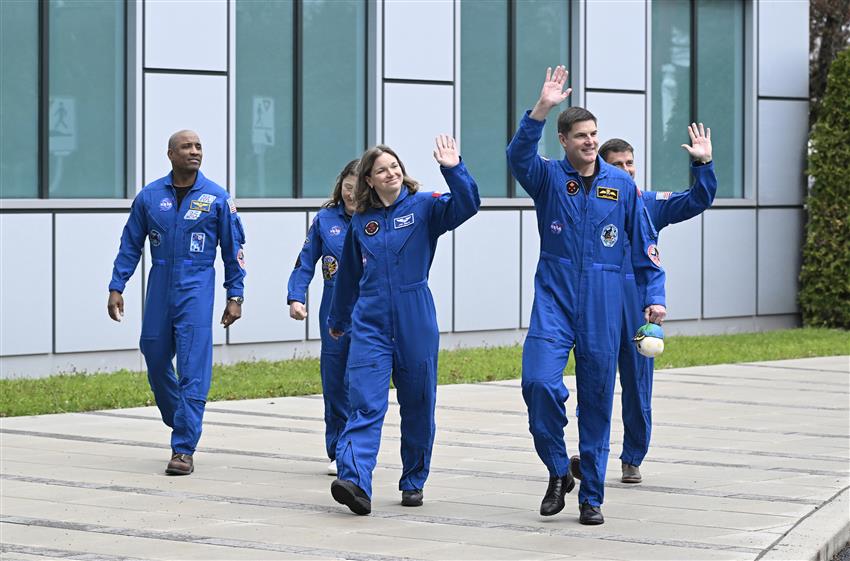 Five astronauts walk in front of the CSA while waving their hands.