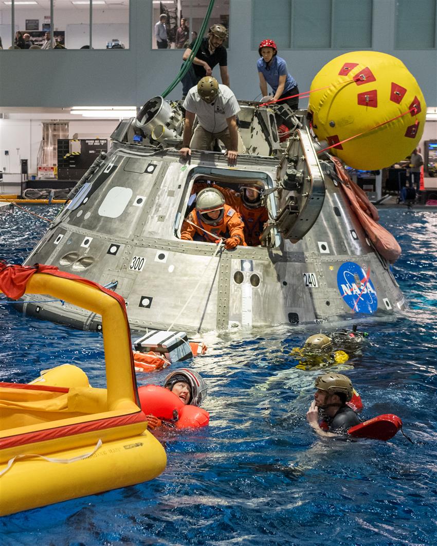 Astronauts in spacesuits and specialized personnel perform a space capsule evacuation drill in a huge indoor pool.