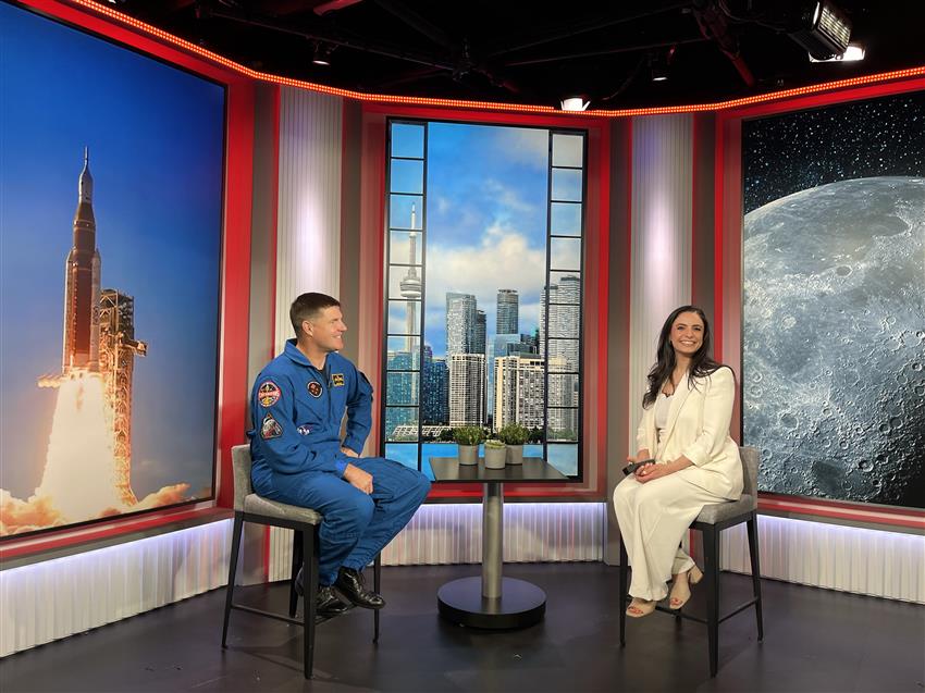 Jeremy and Arda sit on stools with large photos of a rocket, the Toronto landscape and the Moon in the backgro