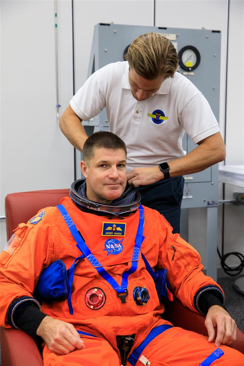 An astronaut is wearing an orange spacesuit, and a man helps him adjust the collar.