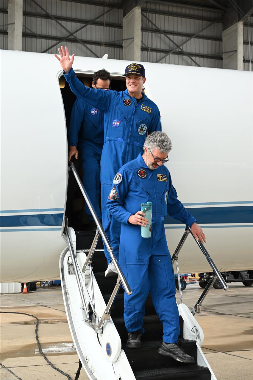 Jeremy waves from the staircase of a plane while Raffi walks in front of him. 