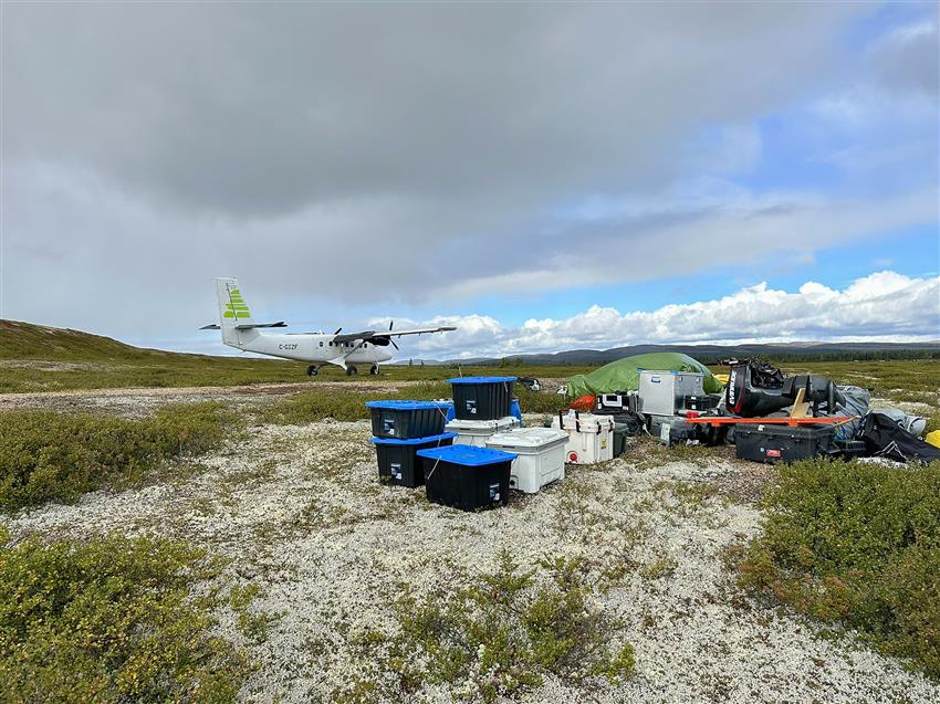 A small plane sits in the tundra. Several boxes and various equipment are in the foreground.