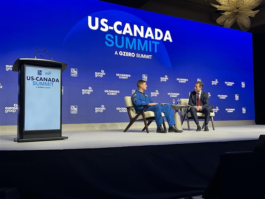 Jeremy and John sit on a stage. Behind them is a big screen that says US–Canada Summit. 