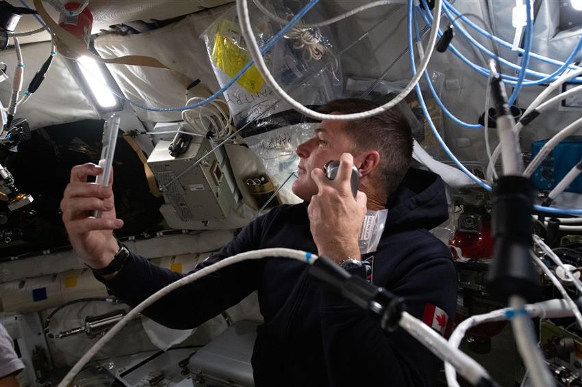 Jeremy shaves inside the Orion spacecraft, holding a phone in one hand and a razor in the other.  