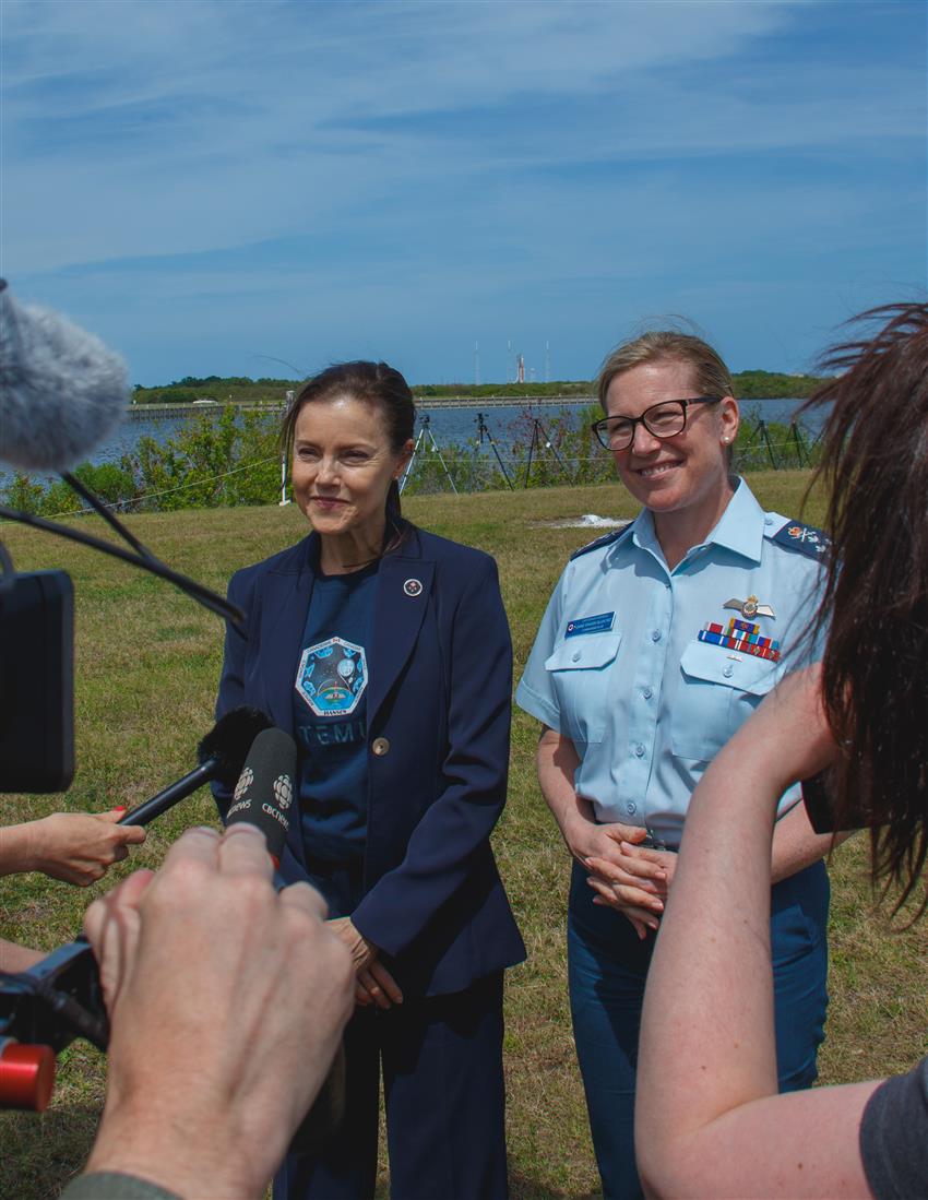 Lisa and Jamie stand in front of journalists with the Artemis II rocket in the distance. 