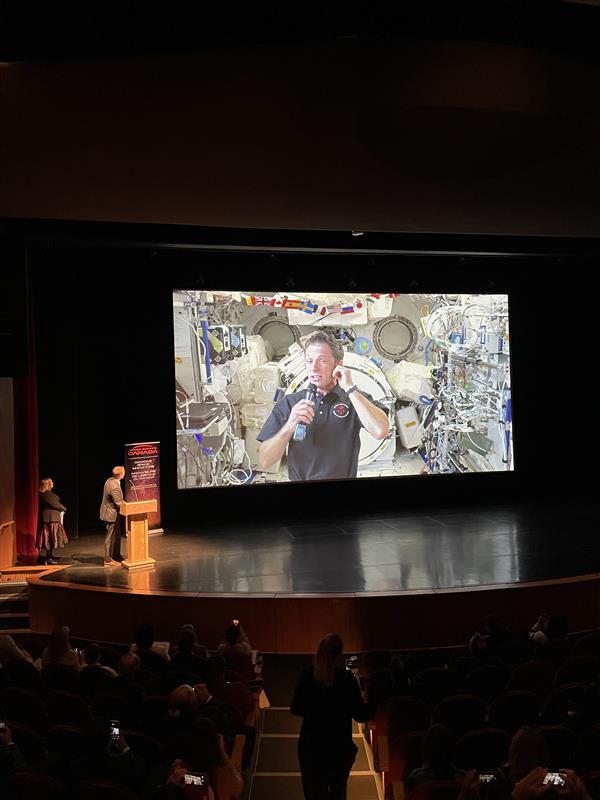 Joshua appears on a screen, speaking live from the Space Station with Brian Gallant, in front of guests.