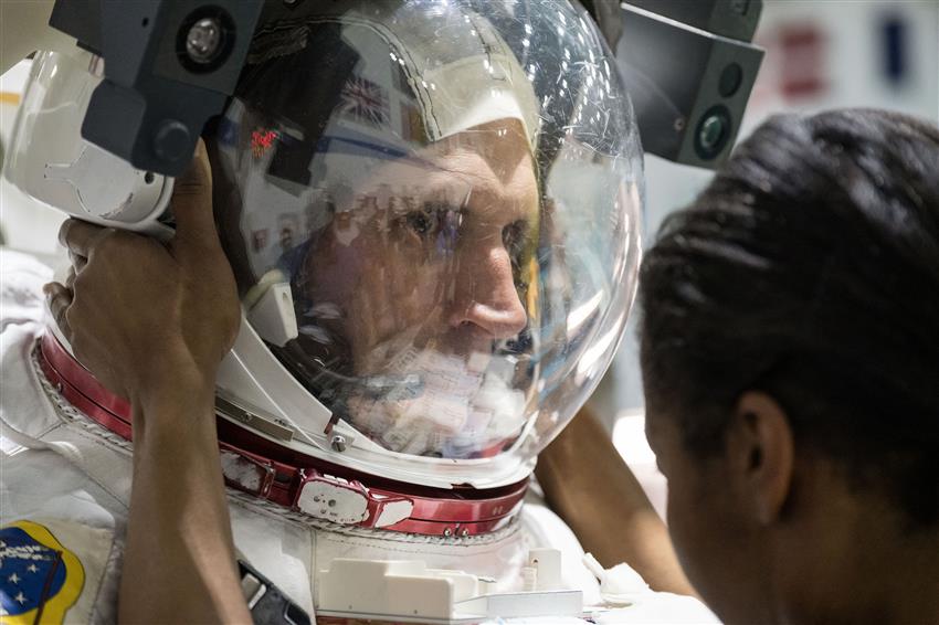 Gros plan de Joshua. Une technicienne finit de lui mettre un casque d'astronaute. 