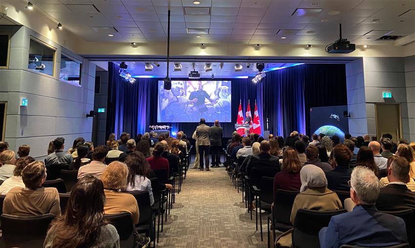 Joshua appears on screen, speaking live from the Space Station, while journalists and guests attend the event.