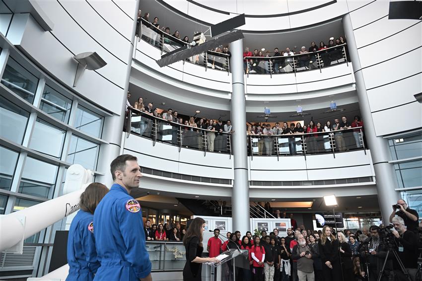 A man and a woman in a blue flight suit face dozens of people assembled on three balconies.