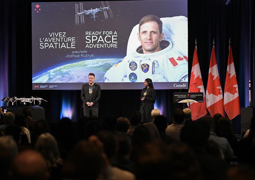 Kumi and Robin are on the stage in front of a large screen where a photograph of Joshua  Kutryk is shown.