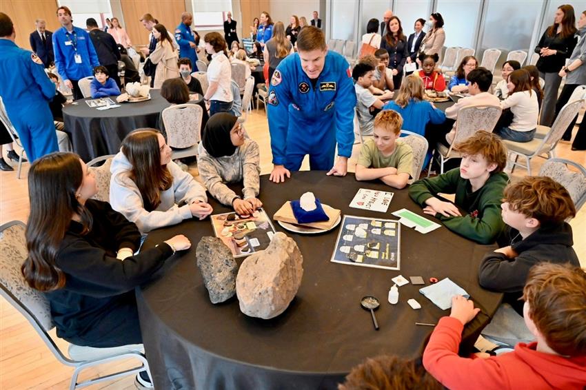 Jeremy speaks with students sitting around a table. Other astronauts are seen around other student tables.