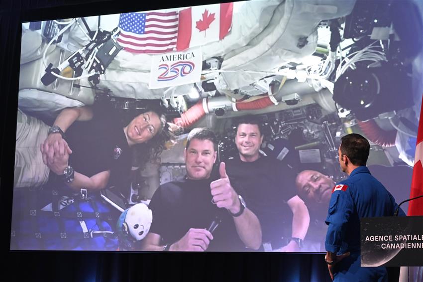 Joshua looks at a big screen with the crew on it. Jeremy is giving a thumbs-up.