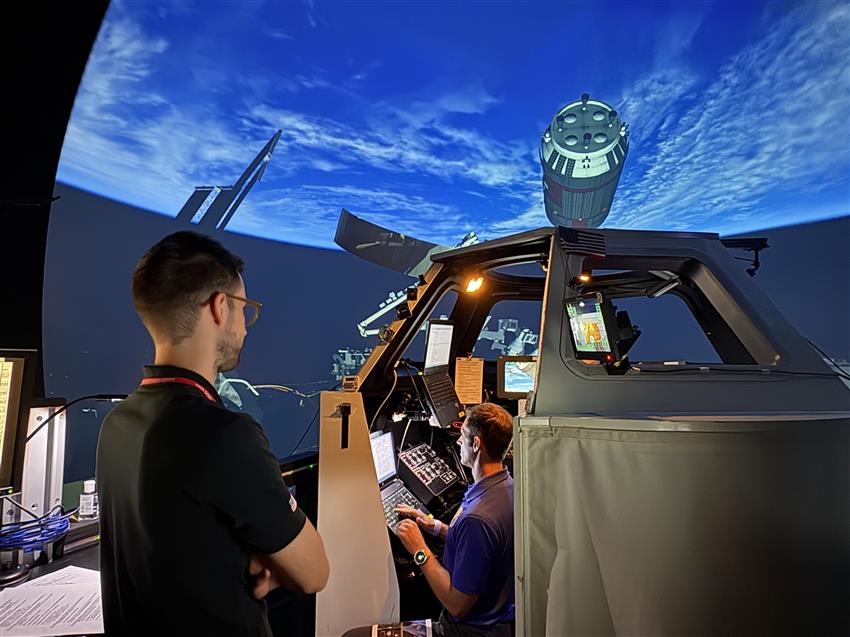 Joshua se trouve dans un simulateur, entouré d'écrans, tandis que Calvin l'observe.