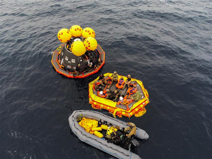 A dozen individuals on two different inflatable boats and a spacecraft mockup floating on the ocean.