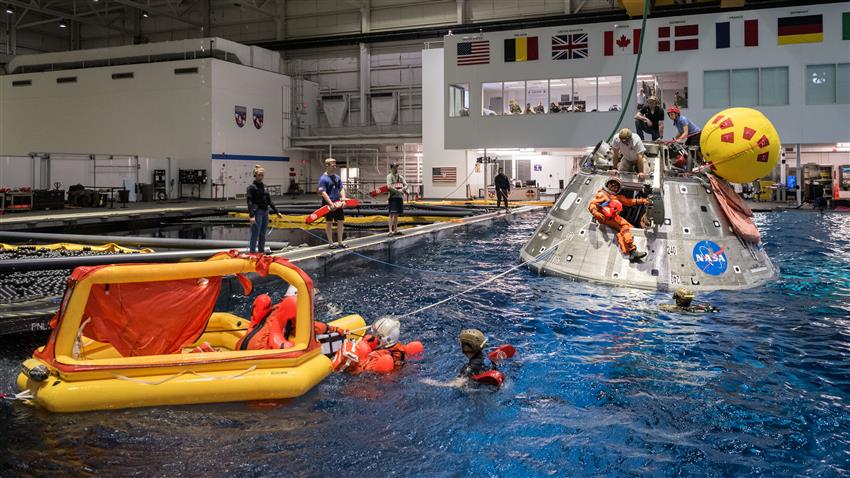 Several people perform a space capsule evacuation drill in a huge indoor pool.