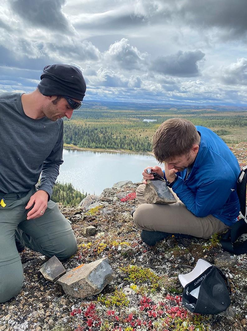 Two men are sitting on the edge of a crater. They are each looking at a rock samples. 
