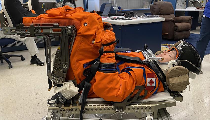 Jenni is wearing an orange spacesuit and is in a seat in which the back is parallel to the ground.