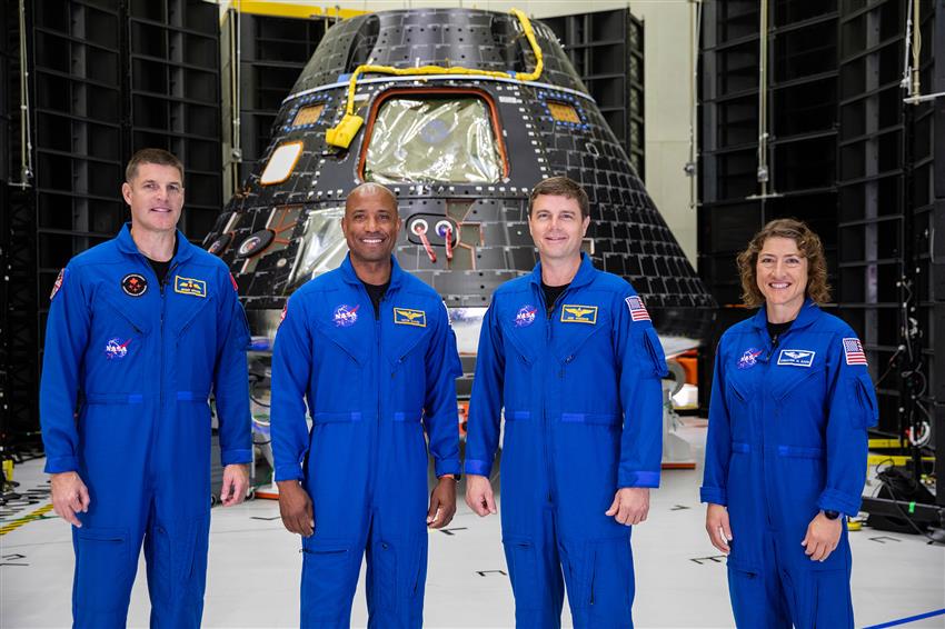 Four astronauts wearing blue flight suits pose in front of a tear-shaped capsule.