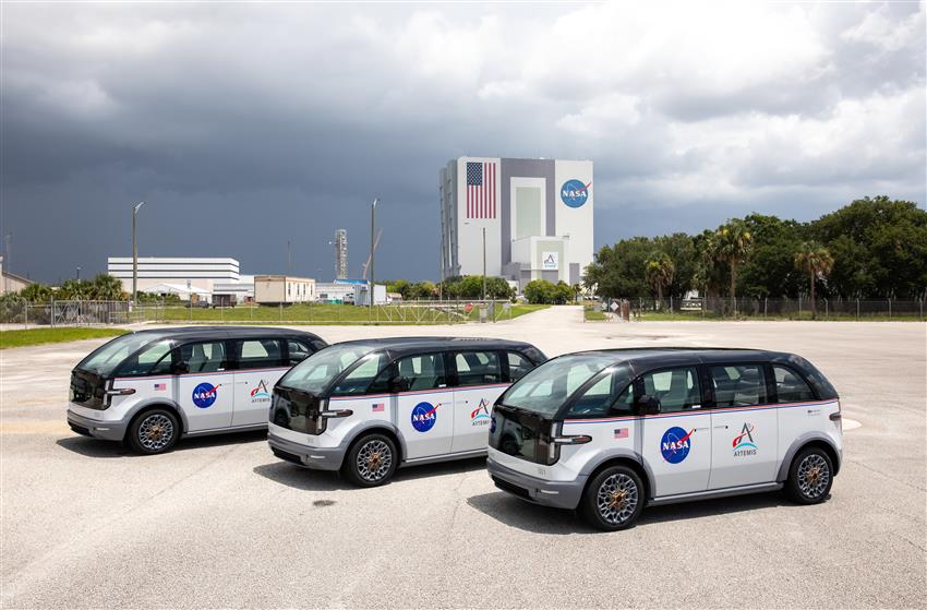 Three van-sized vehicles are parked in a row. NASA's Vehicle Assembly Building is prominent in the background.